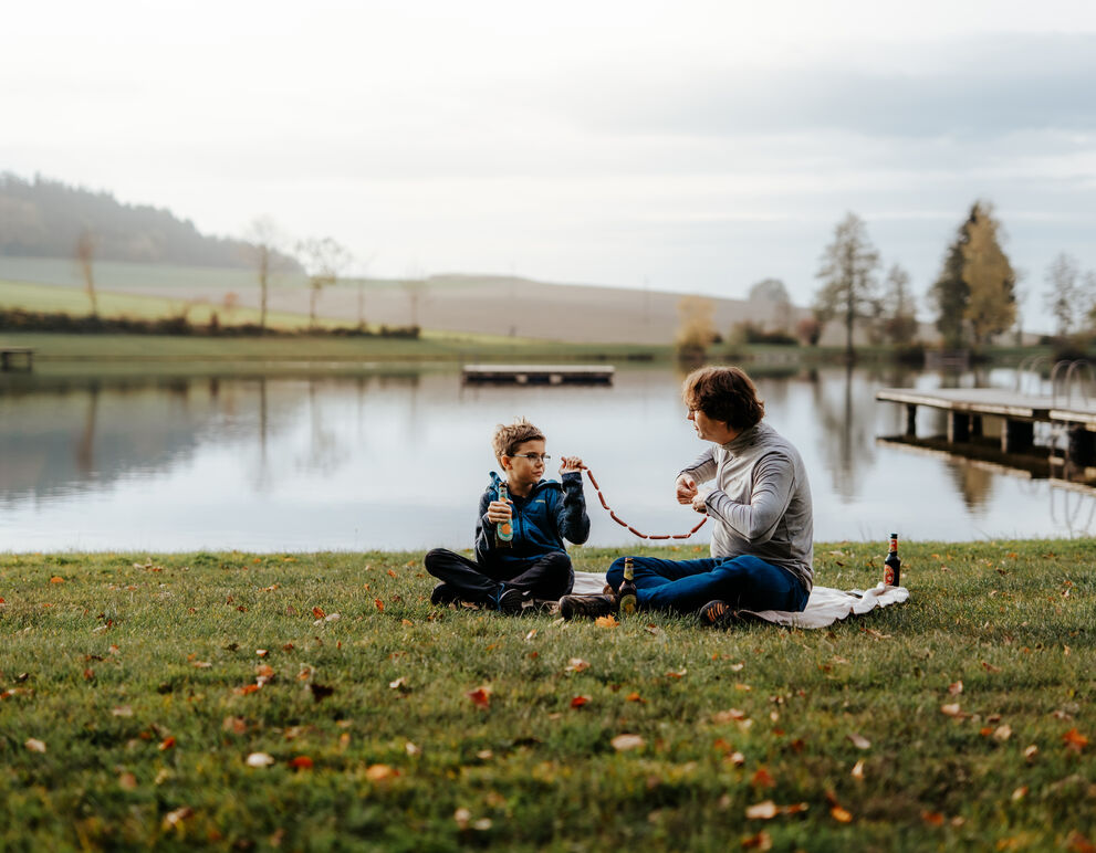 Vater und Sohn sitzen an Ufer eines Badesees und jausenen zusammen;